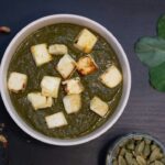 Palak Paneer served in a copper bowl, vibrant green spinach curry with golden paneer cubes, garnished with cream and fresh ginger, overhead shot on a warm wooden surface