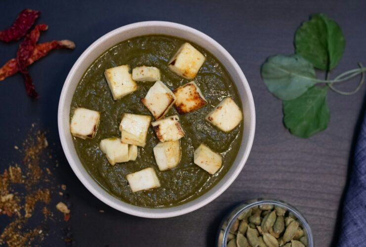 Palak Paneer served in a copper bowl, vibrant green spinach curry with golden paneer cubes, garnished with cream and fresh ginger, overhead shot on a warm wooden surface
