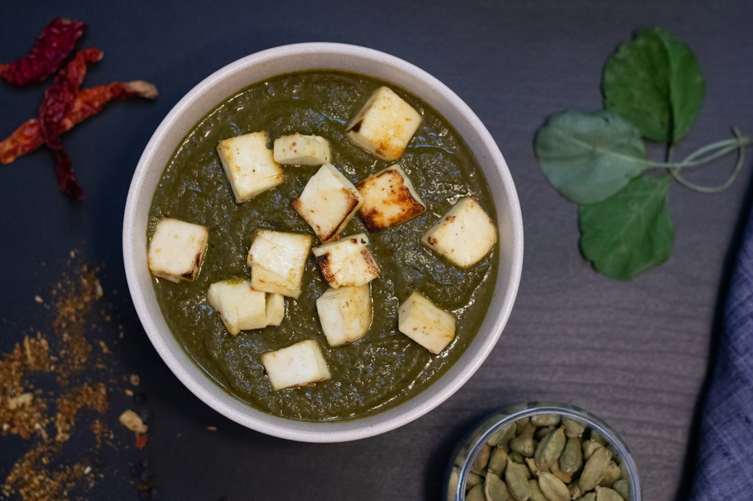 Palak Paneer served in a copper bowl, vibrant green spinach curry with golden paneer cubes, garnished with cream and fresh ginger, overhead shot on a warm wooden surface