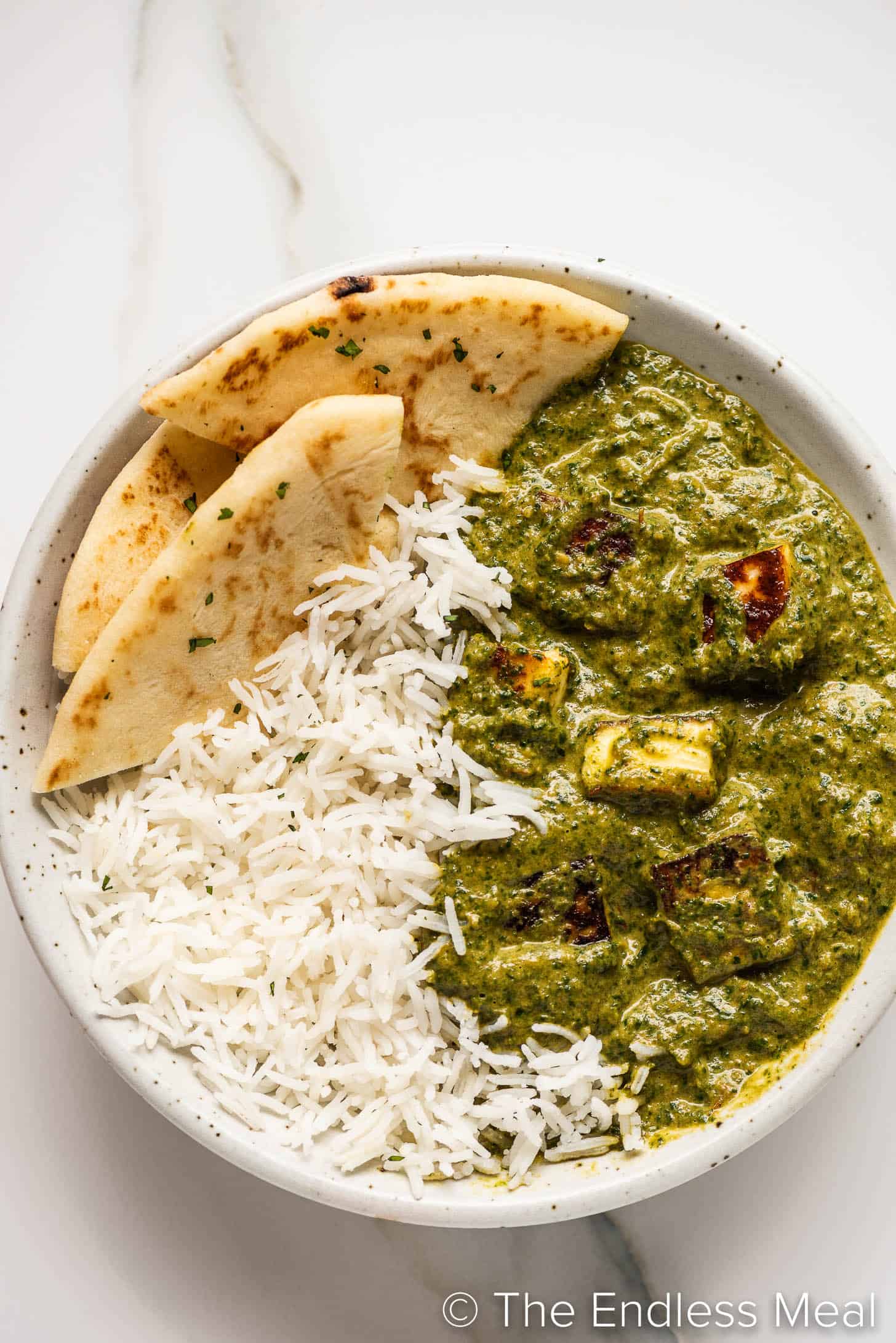 Close-up of Palak Paneer in a bowl with a swirl of cream and a sprinkle of garam masala, golden paneer cubes visible through vibrant green spinach sauce, with a piece of naan resting on the side