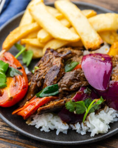 Peruvian Lomo Saltado plated beautifully in a wide bowl with golden french fries, tender beef strips, vibrant tomatoes and red onions, garnished with fresh cilantro