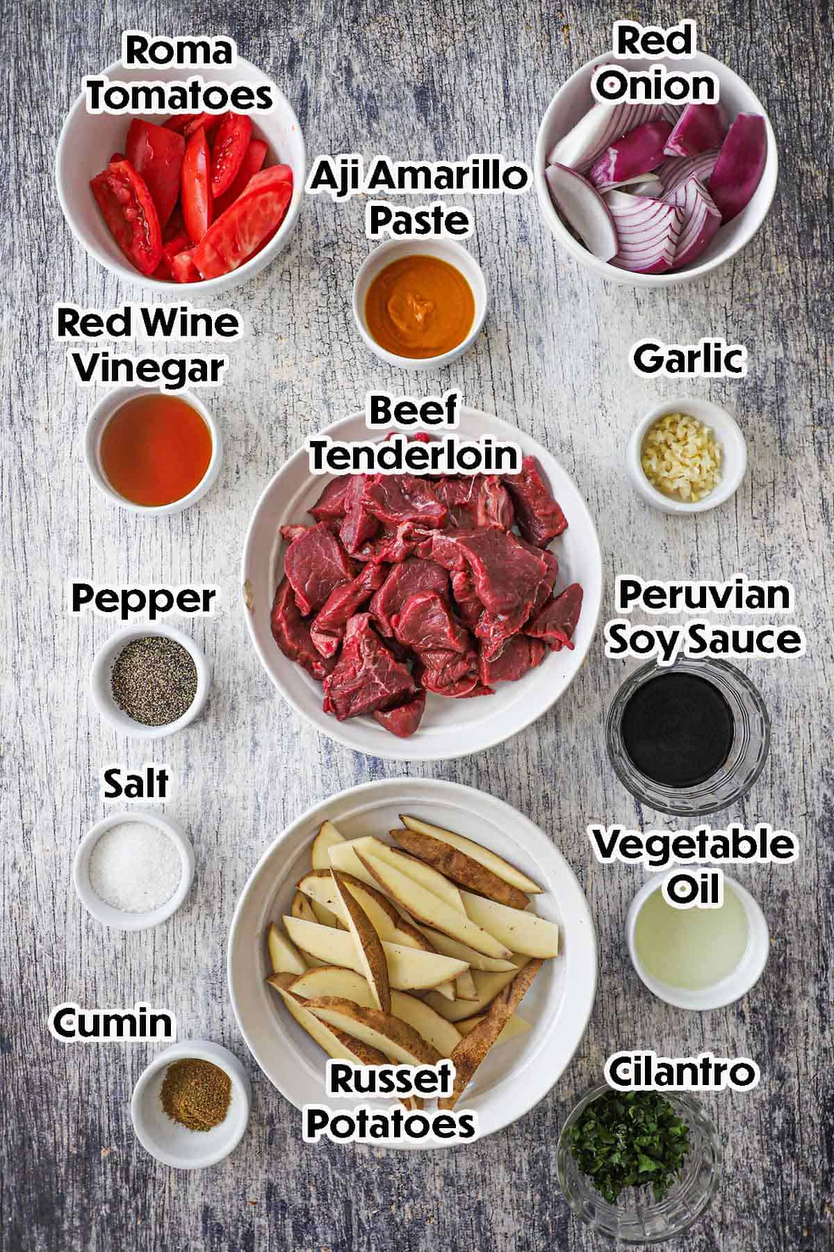 Ingredients for Peruvian Lomo Saltado laid out flat on a wooden board — sliced beef sirloin, roma tomatoes, red onion, aji amarillo paste, soy sauce, and fresh cilantro