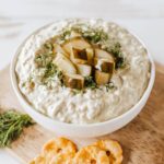 creamy pickle dip in a white bowl garnished with fresh dill and chopped dill pickles, surrounded by crackers and vegetables on a wooden board