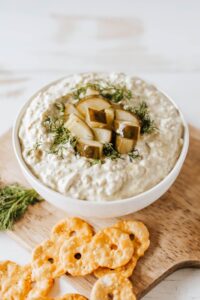 Pickle Dip — Creamy, Tangy, and Completely Addictive 13 creamy pickle dip in a white bowl garnished with fresh dill and chopped dill pickles, surrounded by crackers and vegetables on a wooden board
