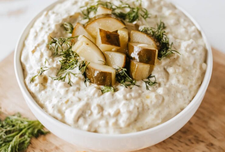 creamy pickle dip in a white bowl garnished with fresh dill and chopped dill pickles, surrounded by crackers and vegetables on a wooden board