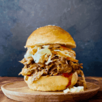 Pulled pork sandwiches piled high on toasted brioche buns with coleslaw and pickles on a rustic wooden board, golden summer light