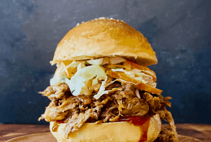 Pulled pork sandwiches piled high on toasted brioche buns with coleslaw and pickles on a rustic wooden board, golden summer light