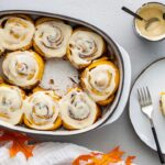 Freshly baked pumpkin cinnamon rolls with cream cheese maple icing drizzled over the top in a white ceramic baking dish, shot from overhead on a rustic wooden table with cinnamon sticks and a small pumpkin as props