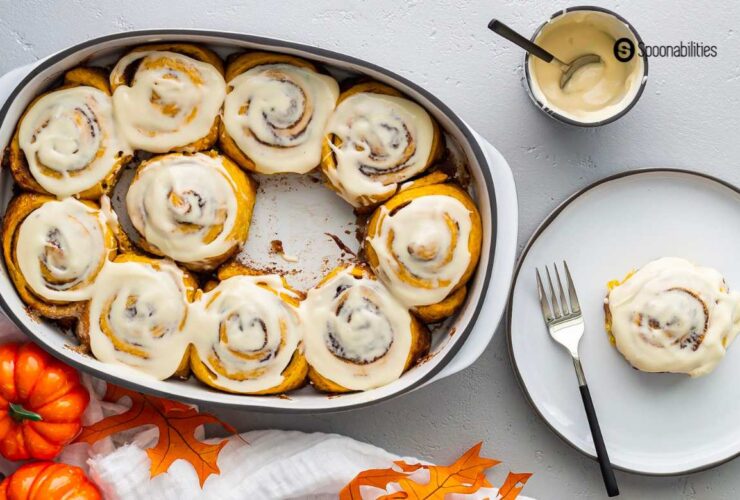 Freshly baked pumpkin cinnamon rolls with cream cheese maple icing drizzled over the top in a white ceramic baking dish, shot from overhead on a rustic wooden table with cinnamon sticks and a small pumpkin as props