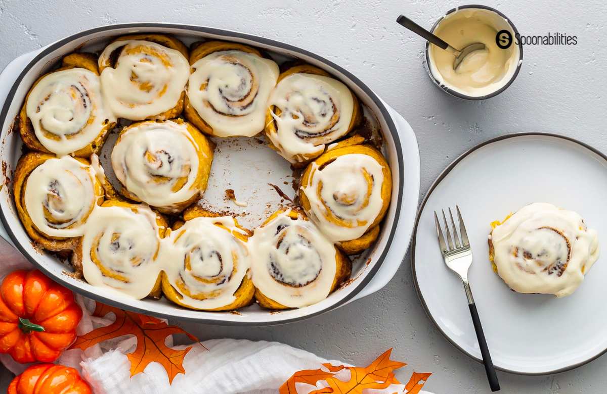 Pumpkin Cinnamon Rolls with Cream Cheese Maple Icing — The Ultimate Overnight Fall Treat 26 Freshly baked pumpkin cinnamon rolls with cream cheese maple icing drizzled over the top in a white ceramic baking dish, shot from overhead on a rustic wooden table with cinnamon sticks and a small pumpkin as props