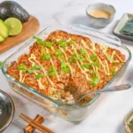 Salmon Sushi Bake served in a white baking dish, golden and bubbly on top, garnished with sliced green onions, tobiko, and drizzled with sriracha and Kewpie mayo, with sheets of nori and cucumber slices on the side