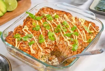 Salmon Sushi Bake served in a white baking dish, golden and bubbly on top, garnished with sliced green onions, tobiko, and drizzled with sriracha and Kewpie mayo, with sheets of nori and cucumber slices on the side