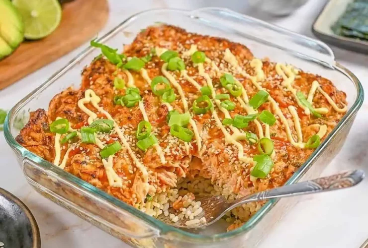 Salmon Sushi Bake served in a white baking dish, golden and bubbly on top, garnished with sliced green onions, tobiko, and drizzled with sriracha and Kewpie mayo, with sheets of nori and cucumber slices on the side