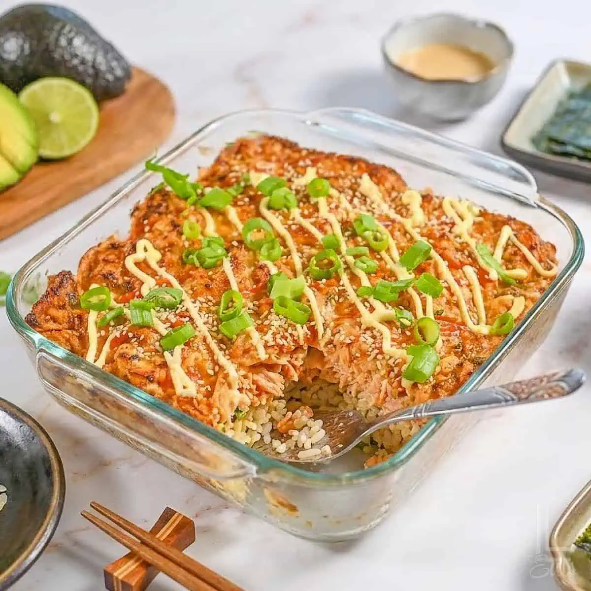 Salmon Sushi Bake served in a white baking dish, golden and bubbly on top, garnished with sliced green onions, tobiko, and drizzled with sriracha and Kewpie mayo, with sheets of nori and cucumber slices on the side