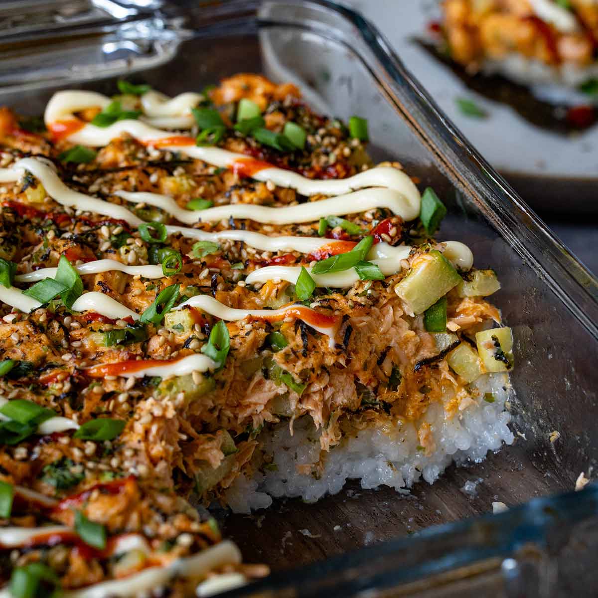 Overhead flat-lay of Salmon Sushi Bake ingredients including raw salmon fillets, a bowl of seasoned sushi rice, Kewpie mayo, cream cheese, sriracha, nori sheets, soy sauce, green onions, and sesame seeds on a light marble surface