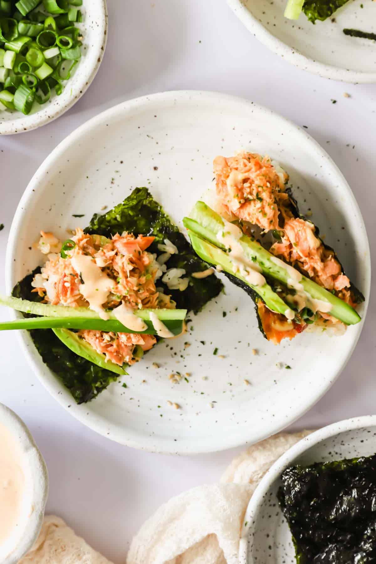 Close-up of a hand scooping a generous portion of creamy salmon sushi bake onto a crispy nori sheet, topped with avocado slices, cucumber, sriracha drizzle, and sesame seeds, ready to eat
