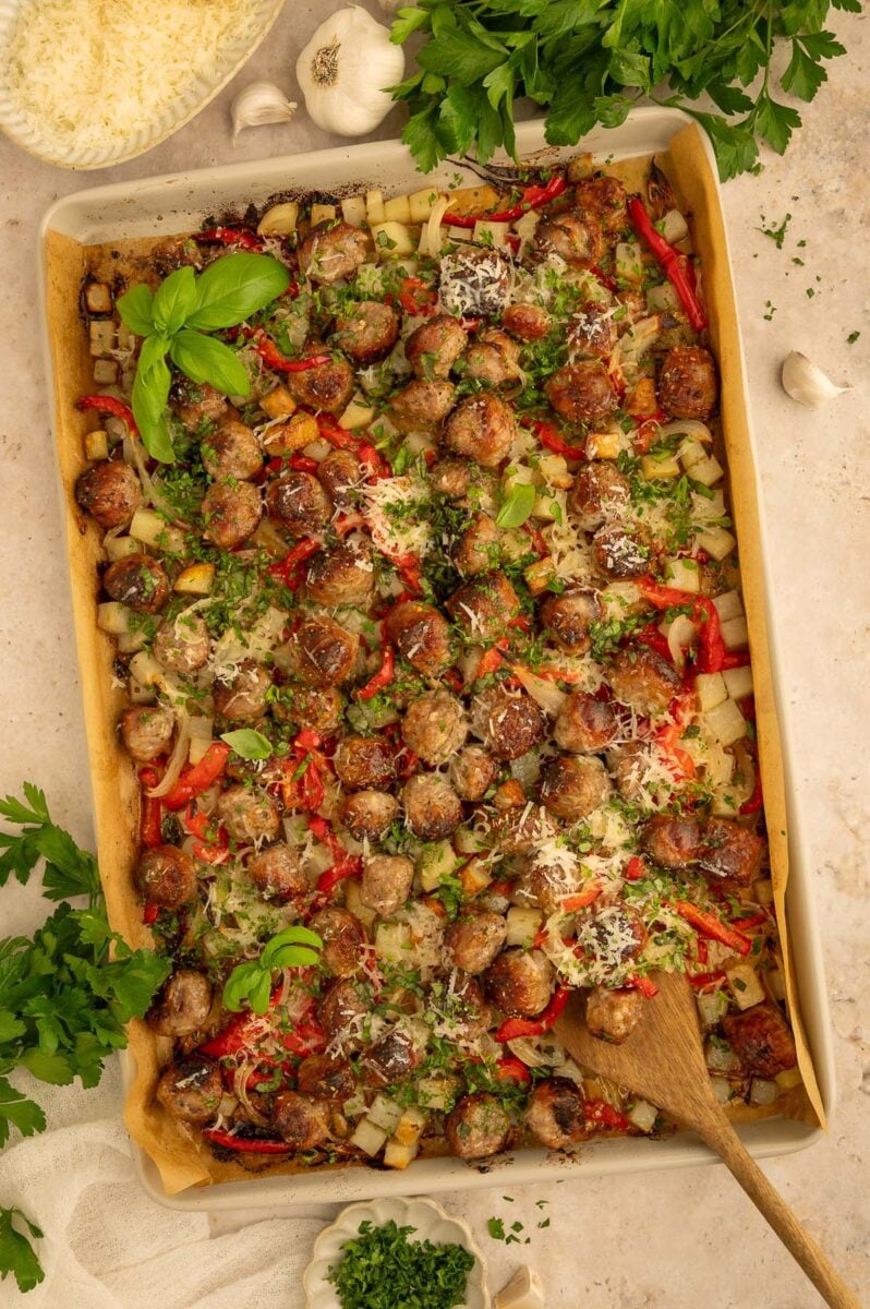 Overhead flat lay of raw ingredients for sheet pan sausage and peppers — sliced bell peppers, halved baby potatoes, onion wedges, raw Italian sausage links, olive oil, and spice jars arranged on a white surface