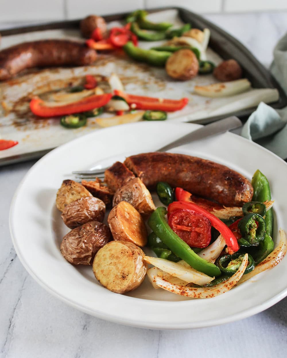 Close-up of sheet pan sausage and peppers served in a white bowl, showing the golden sausage slices, caramelized peppers, and crispy potato edges, with a fork resting on the side