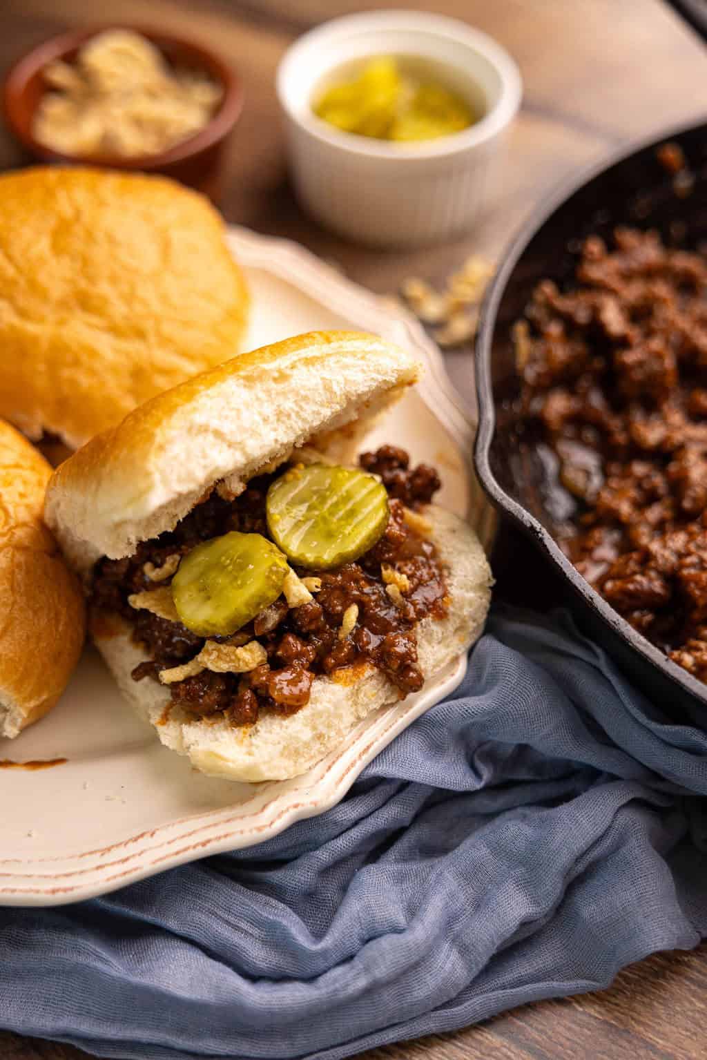 Close-up of a Sloppy Joe sandwich being lifted from a plate, sauce dripping from the toasted brioche bun, with a pickle and chips in the background