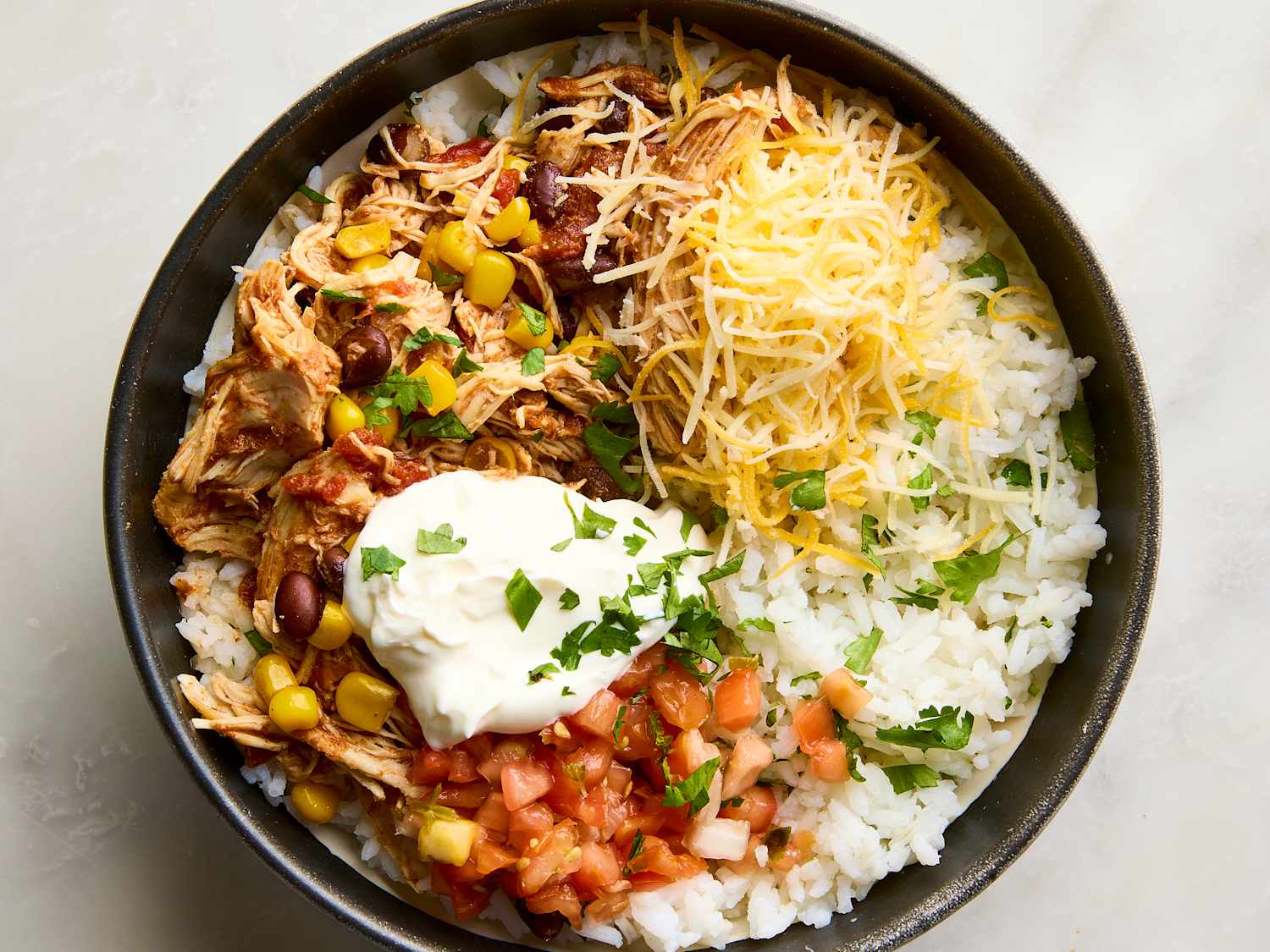 Overhead flat-lay of slow cooker chicken burrito bowl ingredients including raw chicken thighs, canned black beans, corn, enchilada sauce, dry rice, and spices arranged on a white marble surface