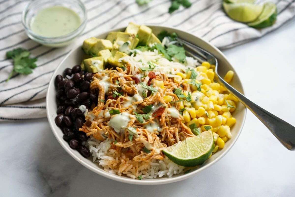Close-up of a slow cooker chicken burrito bowl topped with creamy avocado slices, a dollop of sour cream, fresh cilantro leaves, and a squeeze of lime, photographed from above on a rustic wood surface