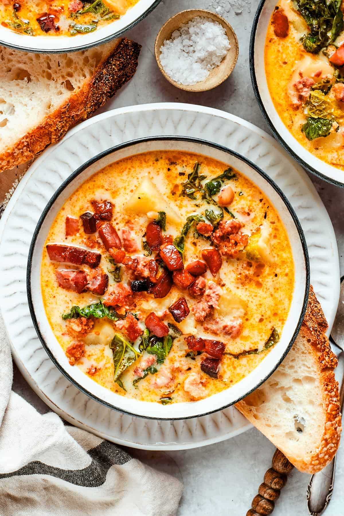 Overhead flat-lay of Slow Cooker Zuppa Toscana ingredients including raw Italian sausage links, sliced Yukon Gold potatoes, fresh kale, garlic cloves, chicken broth carton, and a small pitcher of heavy cream arranged on a rustic wooden surface