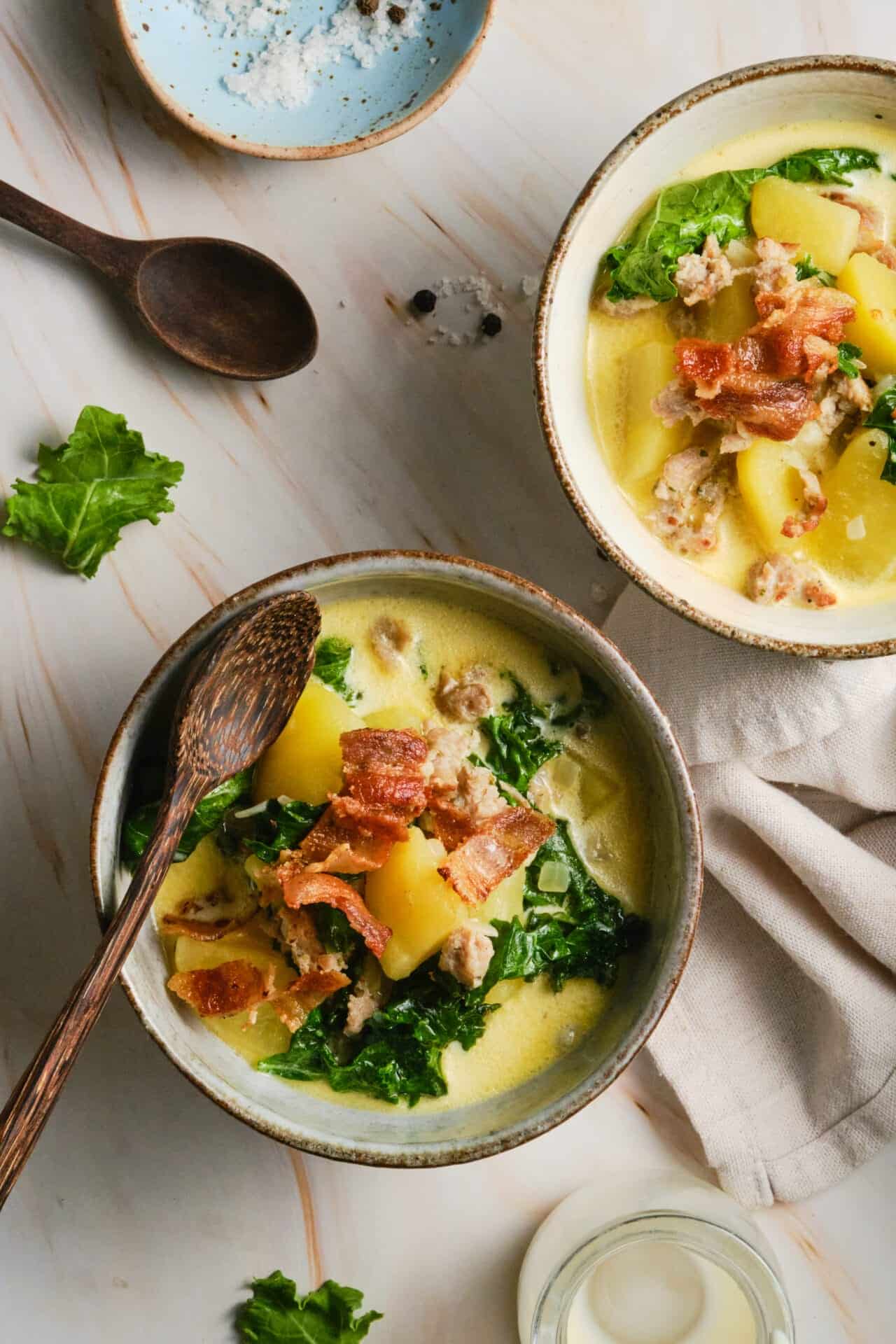 A close-up overhead shot of a rustic bowl of Slow Cooker Zuppa Toscana with visible chunks of potato, slices of sausage, and dark green kale leaves in a pale creamy broth, with a spoon resting on the side and a sprinkle of Parmesan and red pepper flakes on top