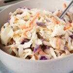 Southern Coleslaw served in a large white bowl with a tangy creamy dressing, garnished with fresh parsley, photographed on a rustic wooden picnic table with BBQ sides in the background