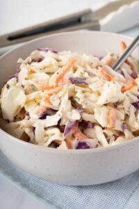 Southern Coleslaw served in a large white bowl with a tangy creamy dressing, garnished with fresh parsley, photographed on a rustic wooden picnic table with BBQ sides in the background