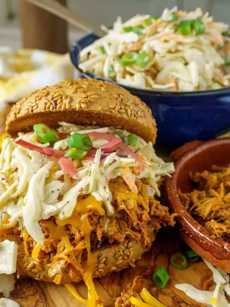 Close-up of Southern Coleslaw piled high on a pulled pork sandwich bun, showing the creamy dressing and vibrant shredded cabbage and carrot colors, with a bowl of extra slaw blurred in the background
