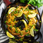 Overhead shot of a vibrant Spanish Paella in a wide pan, golden saffron rice topped with shrimp, mussels, chicken, roasted red peppers, and fresh lemon wedges, photographed on a rustic wooden table