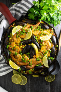 Overhead shot of a vibrant Spanish Paella in a wide pan, golden saffron rice topped with shrimp, mussels, chicken, roasted red peppers, and fresh lemon wedges, photographed on a rustic wooden table