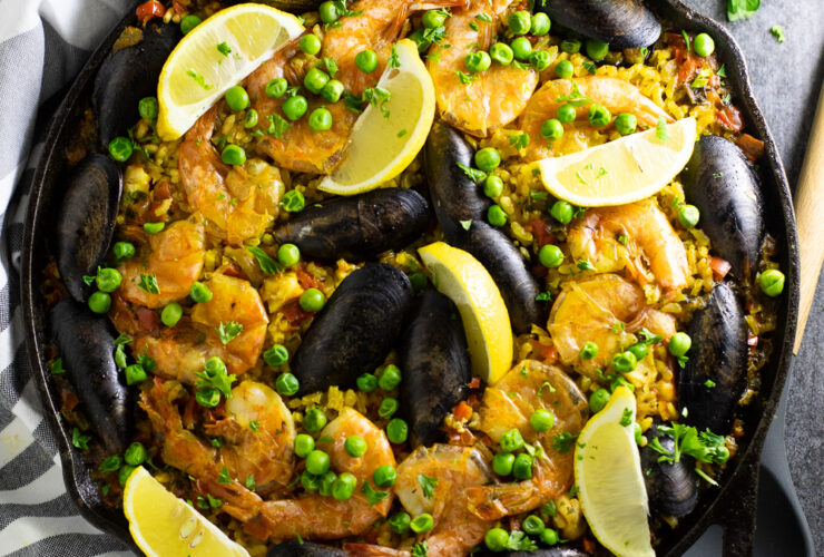 Overhead shot of a vibrant Spanish Paella in a wide pan, golden saffron rice topped with shrimp, mussels, chicken, roasted red peppers, and fresh lemon wedges, photographed on a rustic wooden table