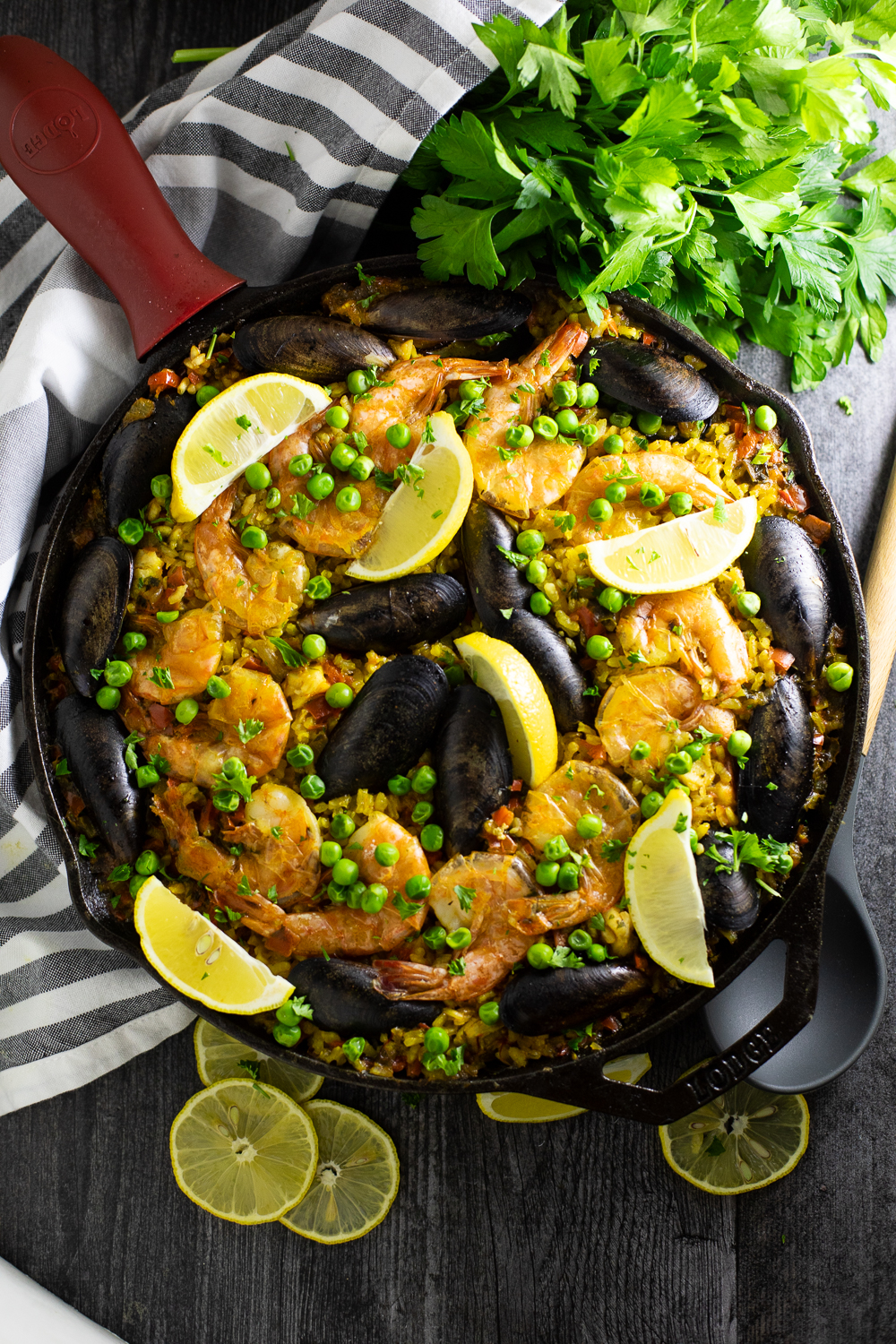 Overhead shot of a vibrant Spanish Paella in a wide pan, golden saffron rice topped with shrimp, mussels, chicken, roasted red peppers, and fresh lemon wedges, photographed on a rustic wooden table