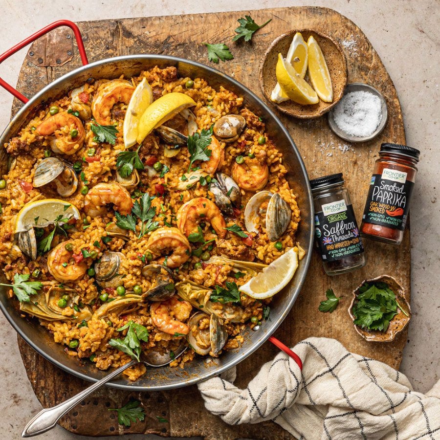 Step-by-step ingredients flat lay for Spanish Paella — raw shrimp, chicken thighs, bomba rice, saffron threads in a small bowl, smoked paprika, roasted red peppers, cherry tomatoes, garlic, and fresh parsley on a marble surface