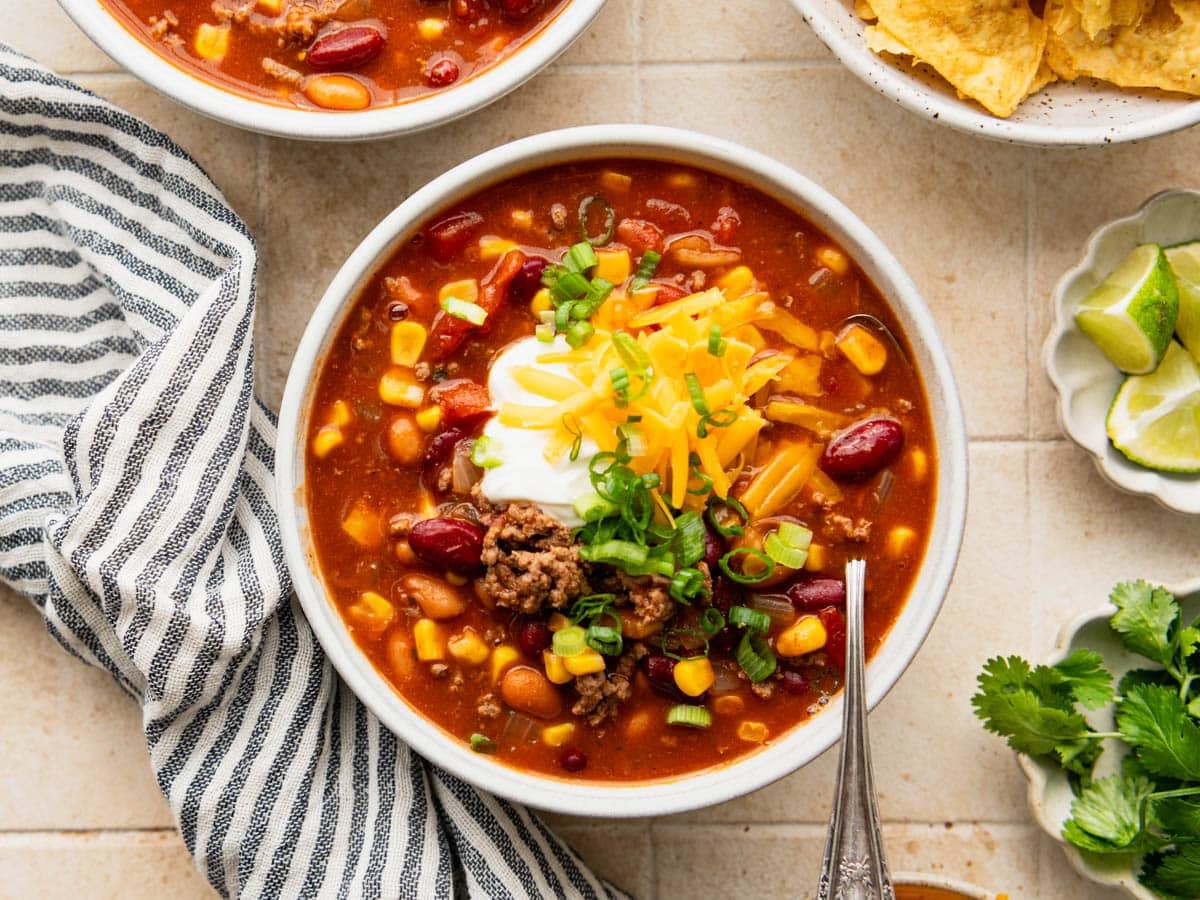 Overhead flat lay of taco soup ingredients including ground beef, canned beans, fire-roasted tomatoes, corn, taco seasoning, broth, and diced onion arranged on a white surface