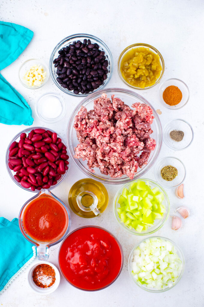 Overhead flat lay of taco soup ingredients including raw ground beef, canned beans, corn, diced tomatoes, taco seasoning, and fresh garnishes arranged on a light marble surface