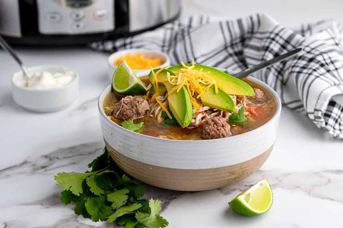 A close-up overhead shot of a bowl of taco soup with vibrant toppings including avocado slices, sour cream swirl, shredded cheese, and fresh lime wedges on the side