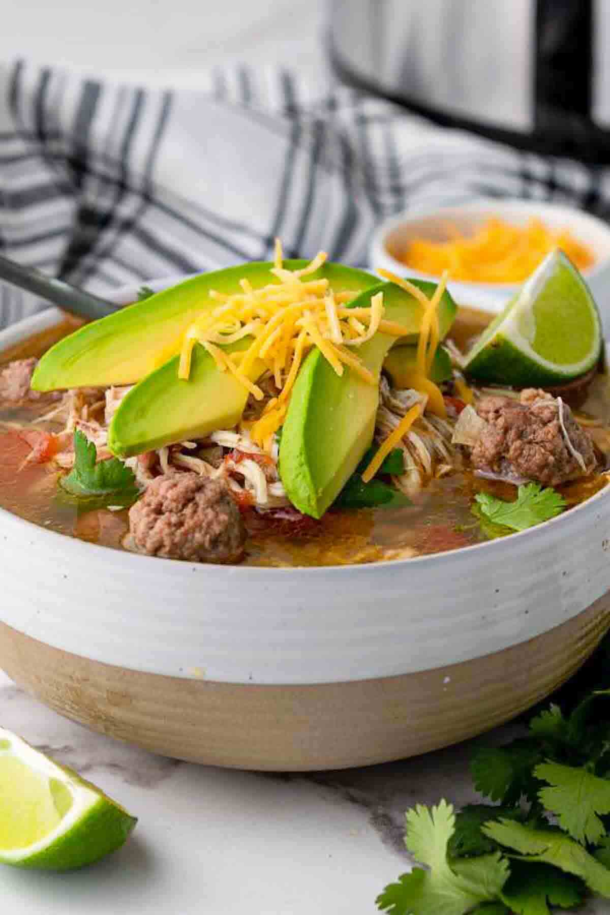 Close-up overhead shot of a bowl of taco soup with vibrant toppings — shredded cheese, avocado slices, sour cream, cilantro, and crushed tortilla chips — with a lime wedge on the side