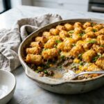 Golden tater tot casserole fresh from the oven in a white baking dish, topped with melted cheddar cheese and crispy tots, garnished with fresh parsley on a rustic wooden table