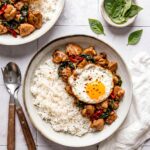 Thai Basil Chicken served over jasmine rice in a white bowl, garnished with fresh basil leaves and a fried egg on top, shot from above on a dark wood surface