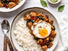 Thai Basil Chicken served over jasmine rice in a white bowl, garnished with fresh basil leaves and a fried egg on top, shot from above on a dark wood surface