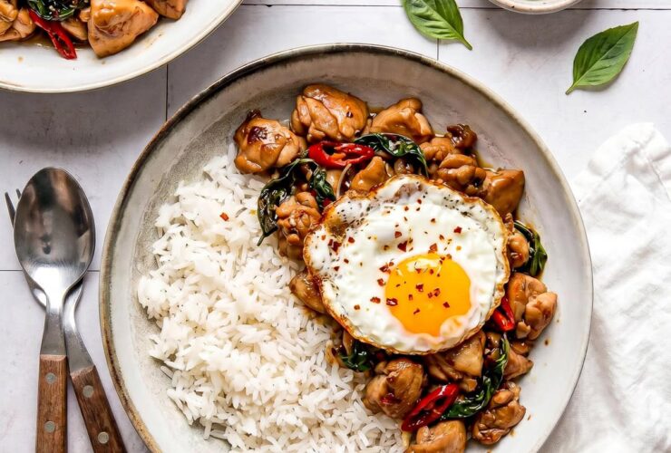 Thai Basil Chicken served over jasmine rice in a white bowl, garnished with fresh basil leaves and a fried egg on top, shot from above on a dark wood surface