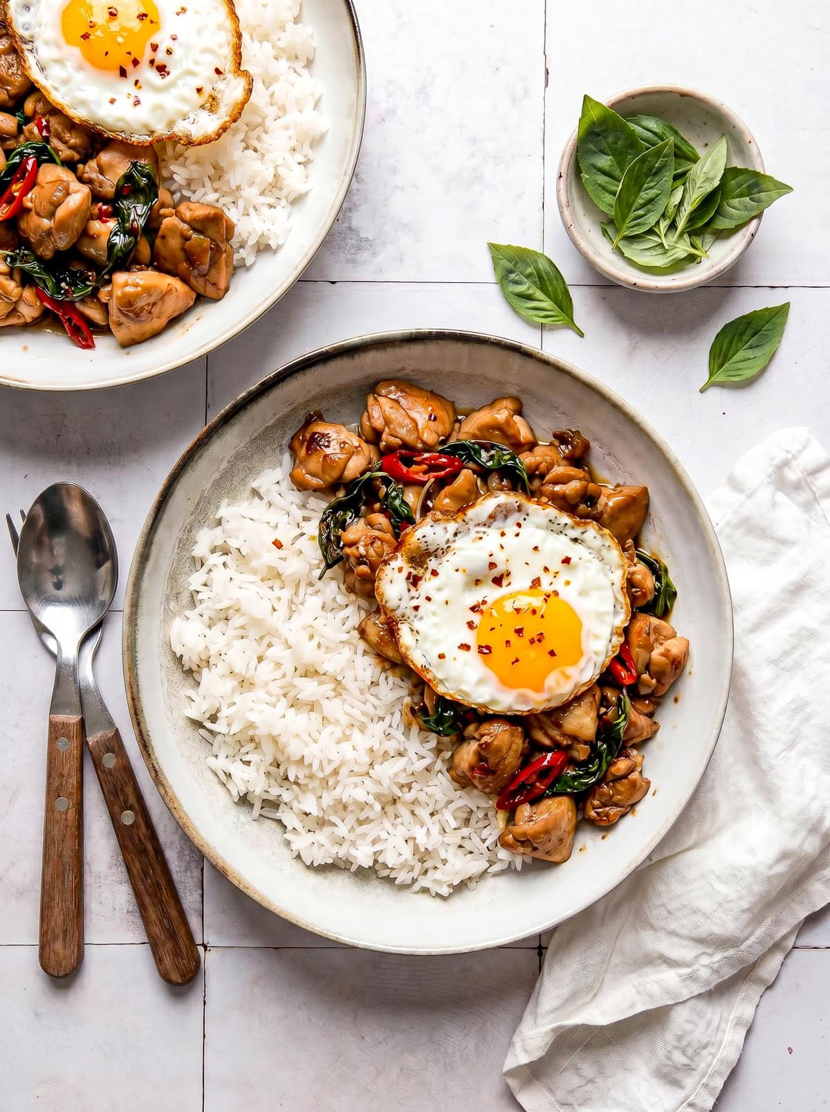 Thai Basil Chicken served over jasmine rice in a white bowl, garnished with fresh basil leaves and a fried egg on top, shot from above on a dark wood surface