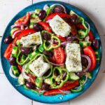 Traditional Greek Salad served on a white ceramic platter with a thick slab of feta, Kalamata olives, chunky tomato wedges, cucumber, green pepper, and red onion drizzled with olive oil and dried oregano
