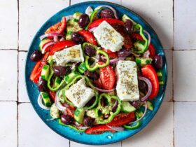 Traditional Greek Salad served on a white ceramic platter with a thick slab of feta, Kalamata olives, chunky tomato wedges, cucumber, green pepper, and red onion drizzled with olive oil and dried oregano