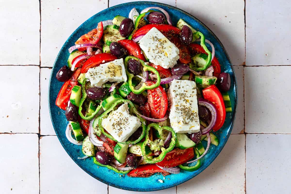 Traditional Greek Salad served on a white ceramic platter with a thick slab of feta, Kalamata olives, chunky tomato wedges, cucumber, green pepper, and red onion drizzled with olive oil and dried oregano