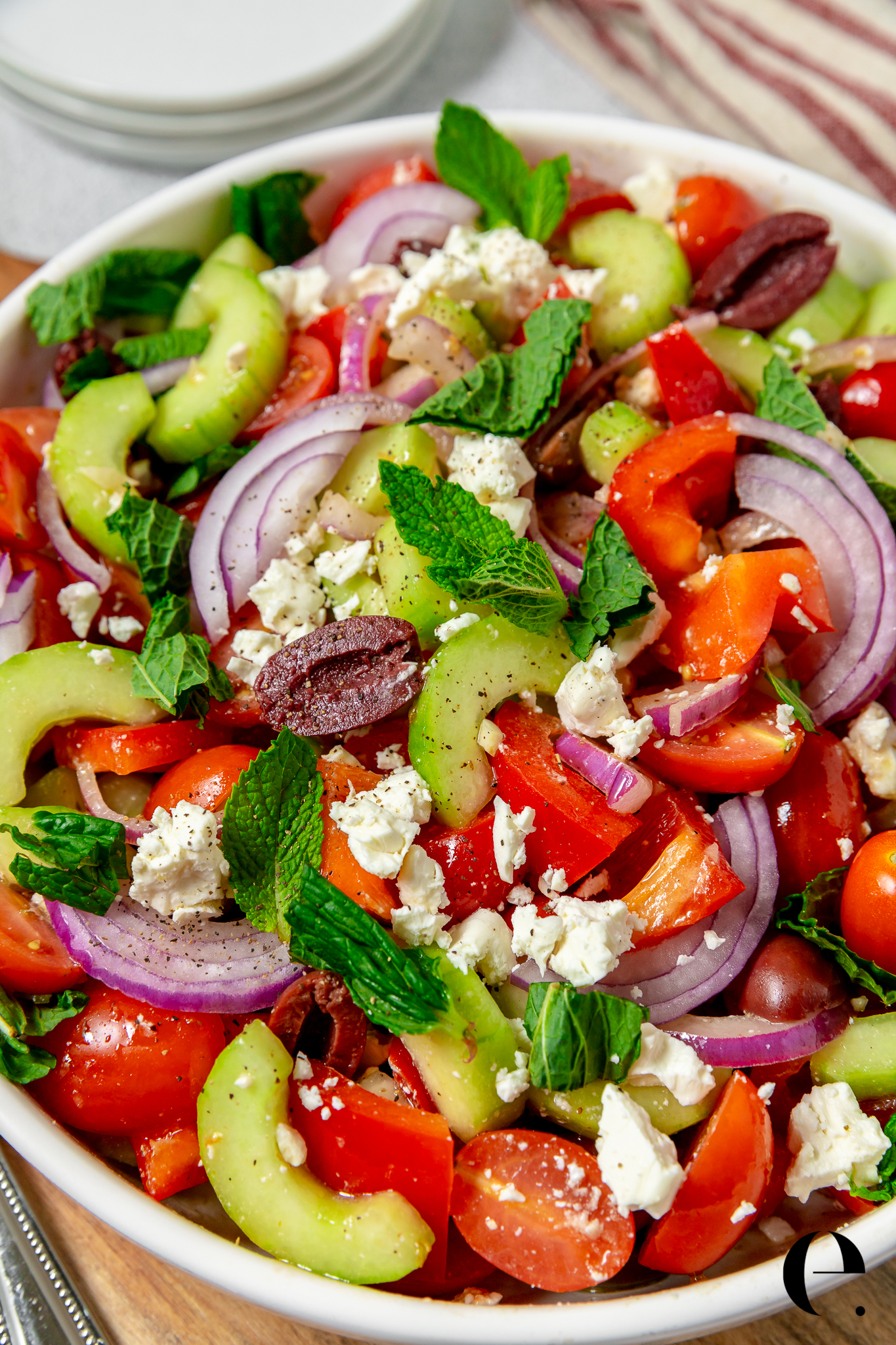 Overhead flat-lay of Traditional Greek Salad ingredients including whole feta block, Kalamata olives, ripe tomatoes, cucumber slices, red onion rings, green pepper strips, and a bottle of extra virgin olive oil on a marble surface