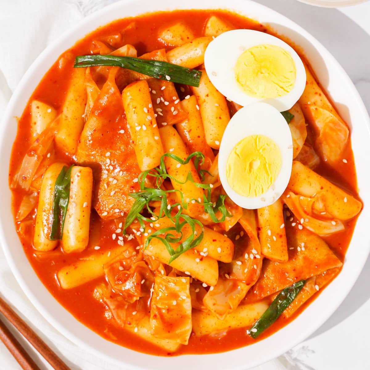 Close-up of tteokbokki rice cakes coated in glossy gochujang sauce with a soft boiled egg halved beside them and sesame seeds scattered on top