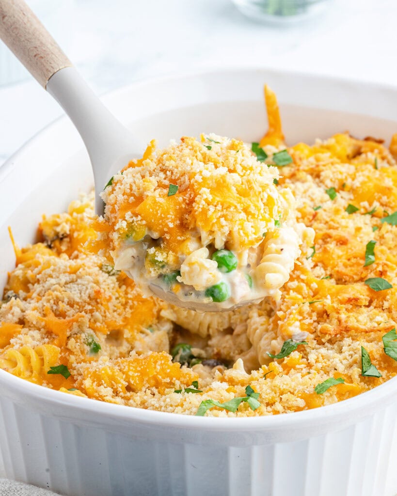 Tuna Casserole — The Classic Comfort Dish Made with Clean, Wholesome Ingredients 28 Close-up of a golden-topped tuna casserole being scooped onto a white plate, showing the creamy noodle and tuna interior with visible peas and melted cheese